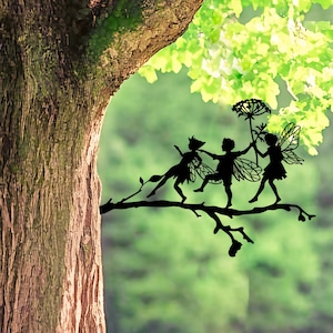Könnte beinhalten: Ein schwarzes Metallsilhouetten-Kunstwerk, das drei Feenfiguren zeigt, die auf einem Ast tanzen. Die Figuren haben zarte Flügel und sind in einer Reihe angeordnet, wobei eine Fee einen hohen, dünnen Blumenstängel mit einer großen, runden Blüte hält.