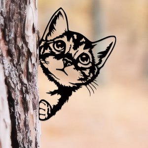 Peut inclure: Une silhouette en métal noir représentant un chaton qui regarde derrière un tronc d'arbre. Le design du chat présente des détails découpés pour les yeux, les oreilles et les moustaches, avec une patte visible près de l'écorce.