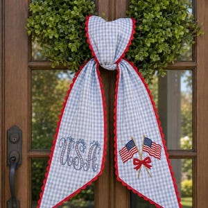 Könnte beinhalten: Ein blau-weißes Gingham-Türschmuck mit roten, gewellten Kanten und einem grünen Kranz. Der Schmuck zeigt die gestickten Buchstaben "USA" und zwei amerikanische Flaggen mit einer roten Schleife.