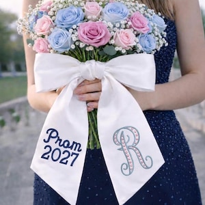 Könnte beinhalten: Ein Strauß aus rosa und blauen Rosen mit weißem Schleierkraut, gebunden mit einer großen weißen Satinschleife. Die Schleife trägt den Text "Prom 2027" und ein monogrammiertes "R" in blauer Stickerei.