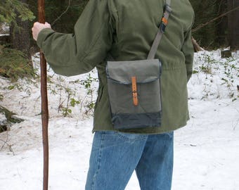 May include: A person wearing a green jacket and blue jeans is carrying a brown leather and canvas messenger bag with a strap over their shoulder. They are holding a wooden walking stick in their left hand.