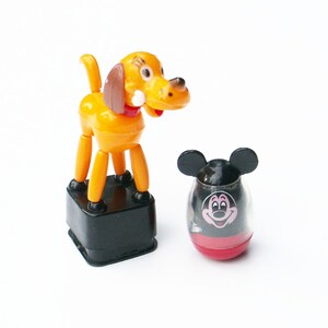 May include: Two vintage Disney toys: a yellow and brown dog figure with a red collar and a black base, and a Mickey Mouse-themed tumbler. The Mickey Mouse tumbler is black with a red base and a smiling Mickey face.