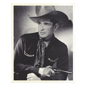 May include: A black and white photo of a man wearing a cowboy hat and a button-down shirt with a bandana around his neck. He is holding a gun in his right hand.