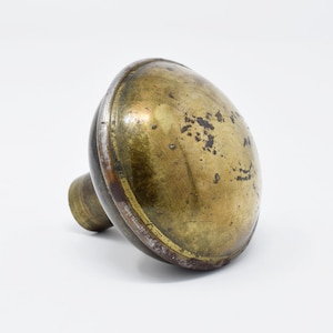 May include: A vintage brass doorknob with a rounded, oval shape. The knob has a tarnished, aged appearance with dark spots and a contrasting ring around the edge. The stem is cylindrical.