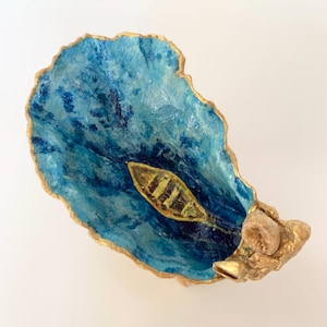 Può includere: Un guscio di ostrica decorativo dipinto con sfumature di blu per assomigliare all'acqua, con una piccola barca gialla al centro. I bordi del guscio sono rifiniti con vernice dorata e un gruppo di piccole conchiglie strutturate è fissato su un lato.