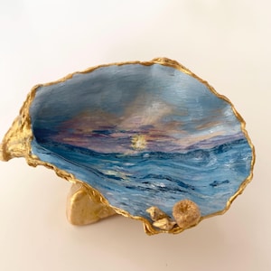 Puede incluir: Una concha de ostra decorativa con un paisaje marino pintado a mano de una puesta de sol sobre olas azules. El interior está pintado en tonos azules, morados y amarillos, con un borde dorado metálico y pequeñas conchas marinas adheridas.