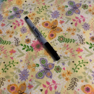 May include: A black Sharpie marker rests on a yellow floral fabric. The fabric features a repeating pattern of butterflies, flowers, and leaves in shades of pink, purple, orange, and green. The background is a soft yellow.