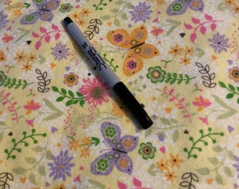 May include: A black Sharpie marker rests on a yellow floral fabric. The fabric features a repeating pattern of butterflies, flowers, and leaves in shades of pink, purple, orange, and green. The background is a soft yellow.