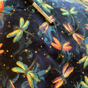 May include: Dark blue fabric with a repeating pattern of colorful dragonflies and gold stars. The dragonflies have wings in shades of orange, pink, yellow, and green. A black marker is visible in the image.