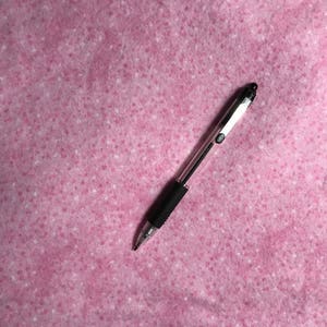 May include: A black and silver ballpoint pen with a black rubber grip rests on a pink, textured surface. The pen is angled diagonally across the frame, showcasing its sleek design and metallic accents. The background is a soft pink with white speckles.