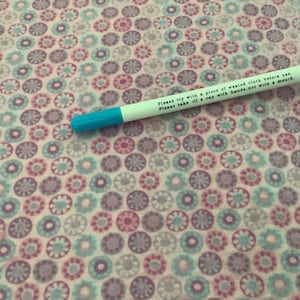 May include: A white pen with a blue tip rests on a fabric patterned with small, circular floral designs in shades of pink, purple, and teal. The pen has text that reads: "Please try with a piece of wasted cloth before use. Please take off a cap with hands, not with a mouth."