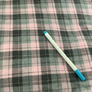 May include: A pink, gray, and white plaid fabric with a white pen with a blue tip. The fabric has a classic checkered pattern, with the pen resting diagonally across the fabric.