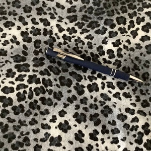 May include: A navy blue pen with silver accents rests on a fabric with a black and gray leopard print pattern. The pen has a silver clip and a silver tip. The fabric has a soft, textured appearance.