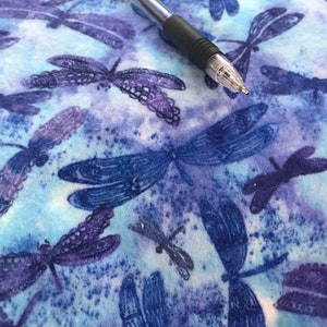 May include: A close-up of a blue and purple dragonfly patterned fabric with a black and silver pen resting on top. The fabric has a watercolour effect with various sizes of dragonflies.