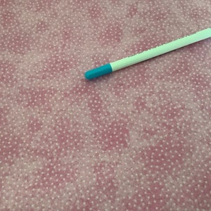 May include: A white and turquoise pen lies on pink fabric with a white polka dot pattern. The pen has a turquoise tip and a white barrel. The fabric has a soft, textured appearance.