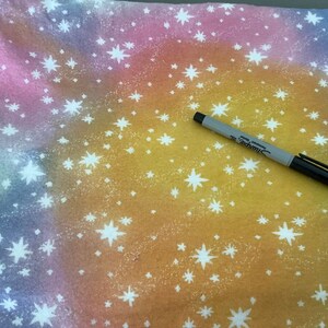 May include: A patterned fabric featuring a celestial design. The background blends from pink to yellow, with white star patterns. A black and grey Sharpie marker is placed on the fabric.