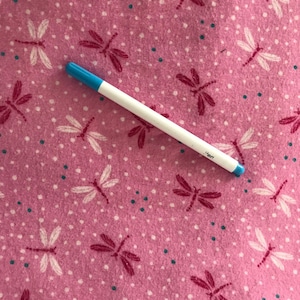 May include: A white pen with a blue tip rests on a pink fabric. The fabric is patterned with white dragonflies, dark pink dragonflies, and small white and teal dots. The pen has the word "pen" printed on it.