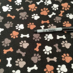 May include: Black fabric with a repeating pattern of white, brown, and gray paw prints and bones. A black Sharpie marker is placed on the fabric.