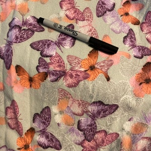 May include: A black Sharpie marker rests on a fabric patterned with butterflies. The butterflies are in shades of orange, purple, and pink, set against a light gray background. The fabric has a soft, textured appearance.