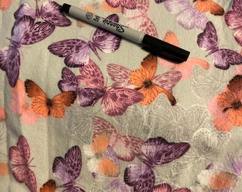 May include: A black Sharpie marker rests on a fabric patterned with butterflies. The butterflies are in shades of orange, purple, and pink, set against a light gray background. The fabric has a soft, textured appearance.