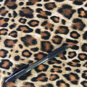 May include: A black pen lies on a leopard print fabric. The fabric has a tan base with brown and black spots, mimicking a leopard's coat. The pen is a standard ballpoint pen, commonly used for writing.