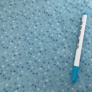 May include: A light blue fabric covered in white and blue stars. A white pen with a blue tip is positioned diagonally across the fabric. The pen has the text "Adger Chaco Ace Pen".