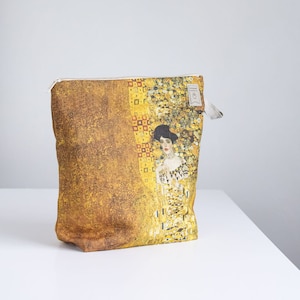 May include: A gold and brown zippered pouch with a painting of a woman in a gold dress. The painting is a reproduction of Gustav Klimt's "The Kiss".
