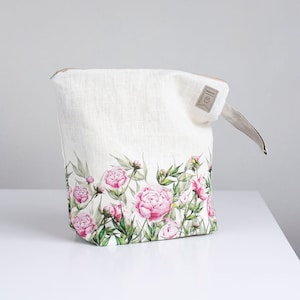 May include: A white linen zippered pouch with a floral print of pink peonies and green leaves. The pouch has a brown leather zipper pull and a small brown leather tag with the word "Handmade" on it.