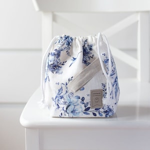 May include: A white drawstring bag with a blue floral pattern and a tan fabric strap. The bag has a small brown tag that says "Handmade".