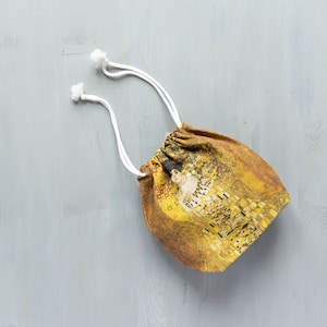 May include: A small drawstring bag with white cords and a printed design of a painting featuring a woman in a gold dress.