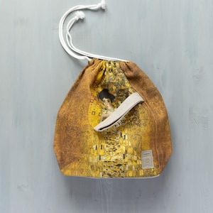 May include: A drawstring bag made of natural fabric with a print of Gustav Klimt's "The Kiss" painting. The bag has a white drawstring and a small brown leather tag with the word "Handmade" on it.