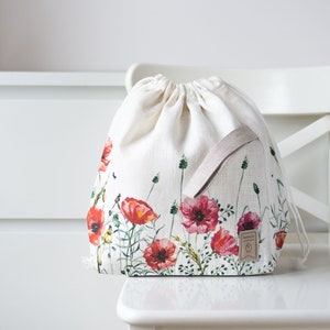 May include: A white drawstring bag with a floral print featuring red poppies and green leaves. The bag has a beige drawstring and a small brown leather tag with the word "Handmade" on it.