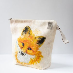 Könnte beinhalten: Beige Leinen-Reißverschlusstasche mit Aquarellmalerei eines Fuchses. Der Fuchs hat orange-gelbes Fell mit schwarzen Akzenten und eine weiße Schnauze. Die Tasche hat eine kleine Stoffschlaufe und ein Etikett mit einem Logo.