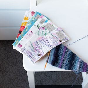 Puede incluir: Cuatro bolsas de tela con estampado floral con un cierre de botón único, una aguja de tejer y un calcetín de punto azul y morado en una silla blanca.