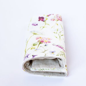 Op de afbeelding: Een witte linnen clutch met een bloemenprint met roze, paarse en groene bloemen. De clutch is dichtgevouwen en heeft een bruine linnen voering.
