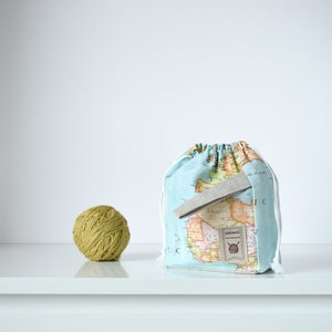 Op de afbeelding: Een lichtblauwe trekkoordtas met een wereldkaartprint. De tas heeft een bruin leren label met het woord "HANDMADE" en een klein afbeelding van een bol garen. Een bol groen garen ligt op een wit oppervlak voor de tas.