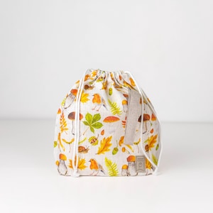Może przedstawiać: Biała torba ze sznurkiem do ściągania, o teksturze lnianej, z kolorowym jesiennym nadrukiem przedstawiającym grzyby, liście, żołędzie i ptaki. Torba ma brązową skórzaną metkę z napisem "Handmade".