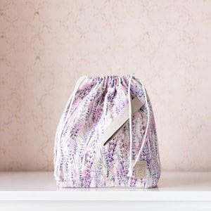 Op de afbeelding: Een witte trekkoordtas met een lavendelbloemenprint en een beige lederen label met de tekst "Handmade".