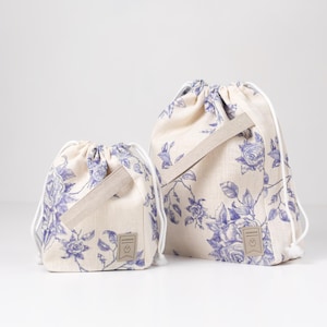 May include: Two drawstring bags with a floral pattern. The bags are made of a light beige fabric with a blue floral print. The larger bag has a brown leather tag with the word "Handmade" on it. The smaller bag has a similar tag.