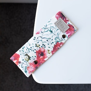Può includere: Un astuccio per matite in tessuto bianco con un motivo floreale con papaveri rossi e foglie verdi. L'astuccio ha un piccolo cartellino in pelle marrone con la scritta "HANDMADE" stampata su di esso.