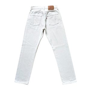 Op de afbeelding: Achterkant van een off-white denim jeans. De jeans heeft een klassiek vijf-zakken ontwerp, een bruine leren patch en een rood label op de achterzak. De jeans is gemaakt van stevig denim.