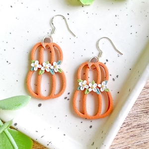 May include: A pair of orange pumpkin-shaped dangle earrings featuring a cutout design. Each earring is decorated with a cluster of three small white flowers with yellow centers and tiny green leaves attached to the center of the pumpkin shape.