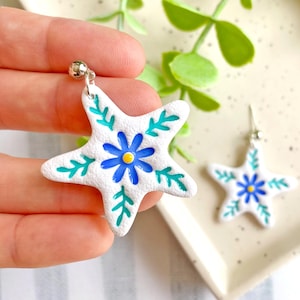 May include: White star-shaped earrings with a blue flower design and green leaf accents. The earrings have a silver-colored hook and a small silver bead at the top. One earring is held in a hand, and the other is on a white tray.