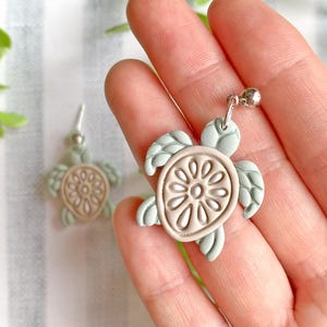 Puede incluir: Pendientes de tortuga verde mar y marrón claro. Los pendientes presentan un diseño de tortuga con caparazón y aletas detalladas. Los pendientes están hechos de un material verde claro con un centro marrón claro. Los pendientes tienen un gancho plateado.