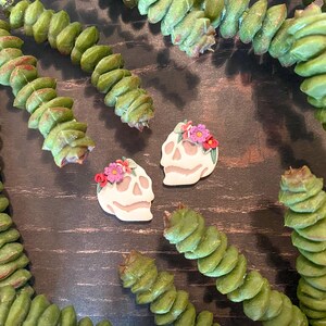 Puede incluir: Dos pendientes con forma de calavera con coronas florales, en crema y rosa, se exhiben sobre una superficie de madera oscura, rodeados de tallos de plantas verdes segmentados. Accesorio artesanal &uacute;nico.