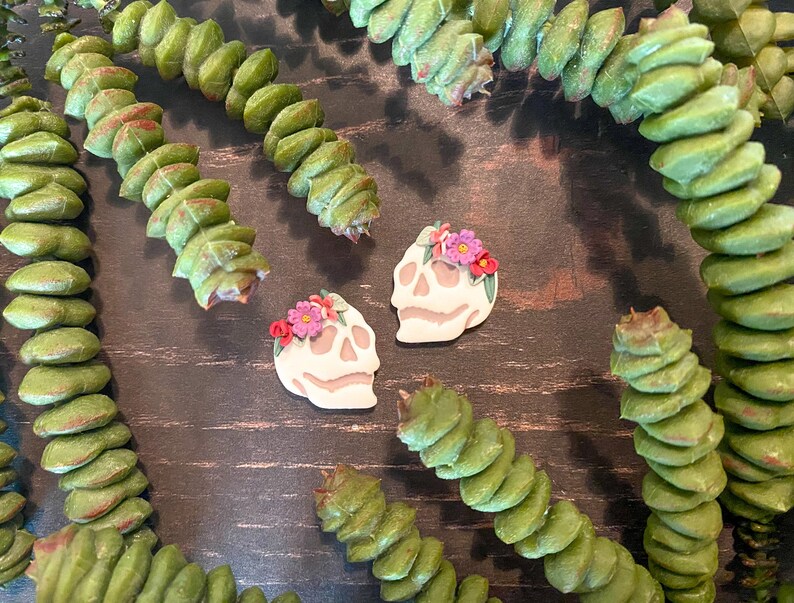 Puede incluir: Dos pendientes con forma de calavera con coronas florales, en crema y rosa, se exhiben sobre una superficie de madera oscura, rodeados de tallos de plantas verdes segmentados. Accesorio artesanal &uacute;nico.