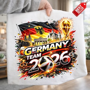 May include: White t-shirt with a colourful graphic promoting the Germany Team 2026. The design incorporates the German flag, a football, a trophy, and the text "GERMANY TEAM 2026". A "Best Seller" tag is attached.