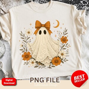 May include: A cream-coloured t-shirt featuring a graphic of a ghost draped in lace fabric with a large orange bow on its head. The ghost is surrounded by orange and white flowers, green leaves, and small gold stars, with a crescent moon in the upper right.