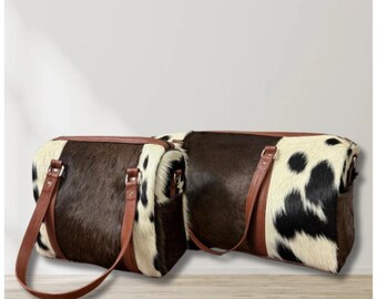 May include: Two cowhide duffel bags. The bags have a brown, black, and white spotted pattern. They feature brown leather trim, handles, and adjustable shoulder straps. The bags are rectangular with rounded edges.
