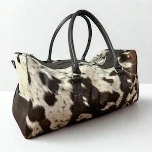 May include: A cowhide leather duffel bag with a spotted pattern in shades of brown and white. The bag features dark brown leather handles and trim, and a zipper closure. This travel bag is suitable for weekend getaways.